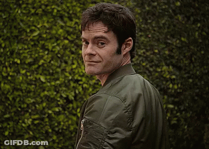 A Man Wearing A Green Bomber Jacket Is Standing In Front Of A Bush . GIF