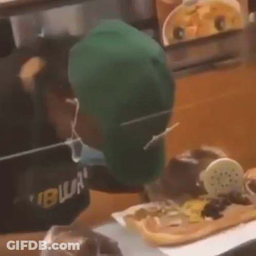 A Man Wearing A Green Hat And A Mask Is Eating A Sandwich . GIF