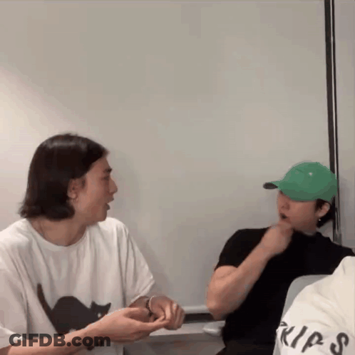 A Man Wearing A Green Hat Is Sitting Next To Another Man Wearing A White Shirt With A Black Cat On It . GIF