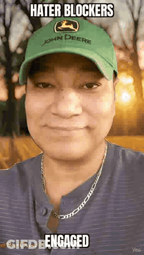 A Man Wearing A Green John Deere Hat And A Necklace Is Smiling And Looking At The Camera . GIF