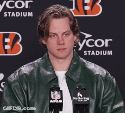 A Man Wearing A Green Leather Jacket Stands In Front Of Two Microphones With A Nfl Logo On Them GIF