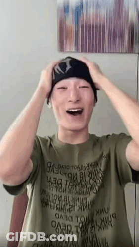 A Man Wearing A Green Shirt And A Black Beanie Is Laughing While Holding His Head . GIF