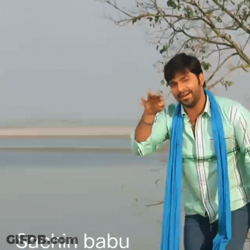 A Man Wearing A Green Shirt And A Blue Scarf Stands In Front Of A Body Of Water GIF