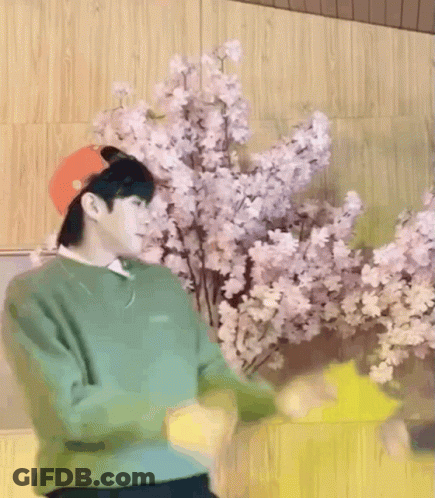A Man Wearing A Green Sweater And A Red Hat Is Dancing In Front Of Flowers GIF