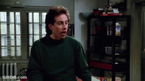 A Man Wearing A Green Sweater Is Sitting In Front Of A Shelf With A Box That Says Dominoes GIF