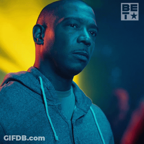 A Man Wearing A Grey Hoodie Stands In Front Of A Sign That Says Be T GIF