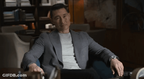 A Man Wearing A Grey Jacket And A Watch Sits In A Dark Room GIF