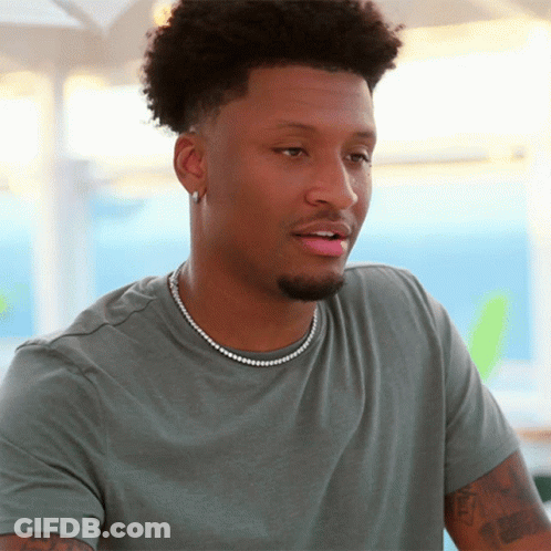 A Man Wearing A Grey Shirt And A Pearl Necklace Looks At The Camera GIF