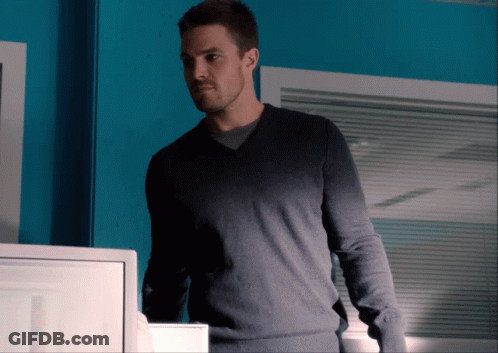 A Man Wearing A Grey Sweater Stands In Front Of A Blue Wall GIF