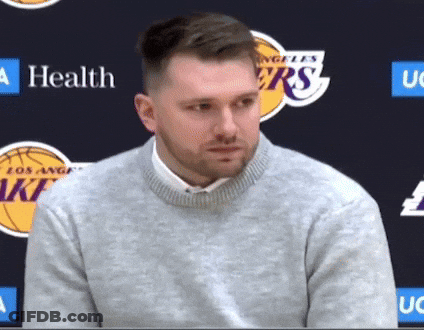 A Man Wearing A Grey Sweater Stands In Front Of A Los Angeles Lakers Logo GIF