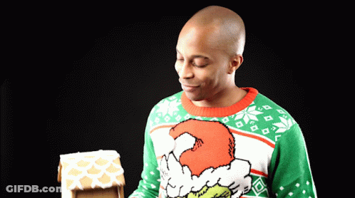A Man Wearing A Grinch Sweater Looks At A Gingerbread House GIF