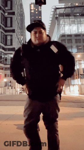 A Man Wearing A Hat And A Black Jacket Stands In Front Of A Tall Building GIF