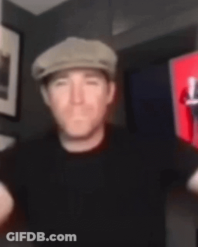 A Man Wearing A Hat And A Black Shirt Is Standing In A Room . GIF