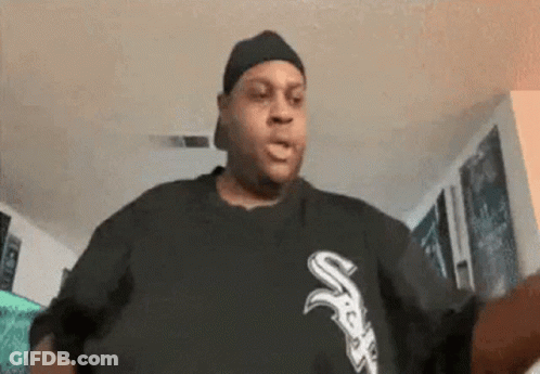 A Man Wearing A Hat And A Black Shirt With The Letter S On It Is Standing In A Living Room . GIF