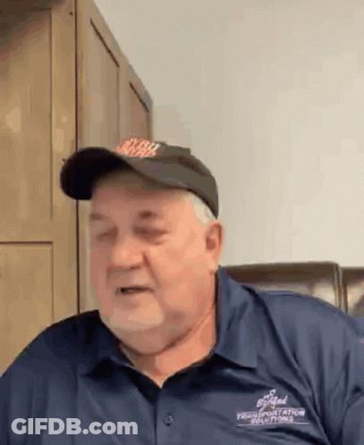 A Man Wearing A Hat And A Blue Shirt Is Sitting In A Chair And Making A Funny Face . GIF