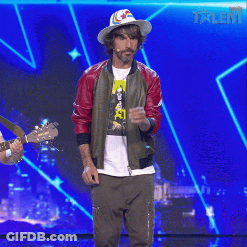 A Man Wearing A Hat And A Bomber Jacket Stands On A Stage With A Blue Background That Says Talent GIF