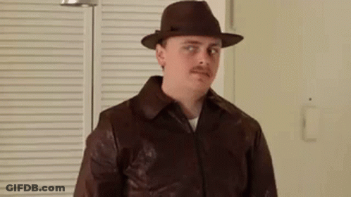 A Man Wearing A Hat And A Brown Leather Jacket Is Standing In Front Of A Window . GIF