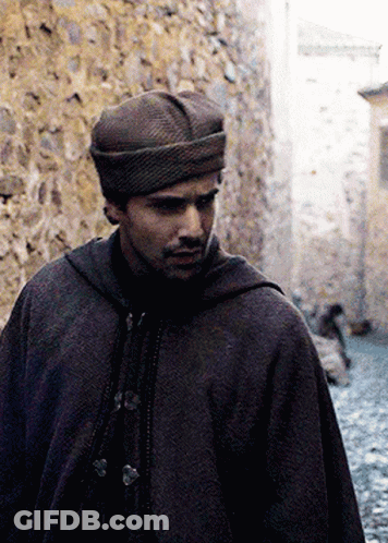 A Man Wearing A Hat And A Cape Is Walking Down A Narrow Street GIF