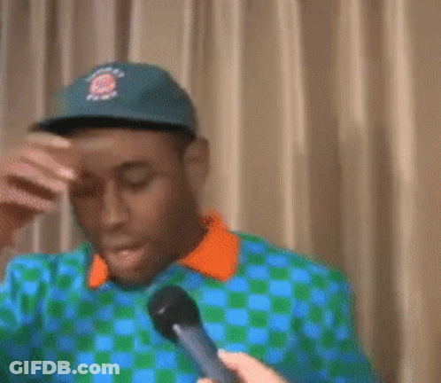 A Man Wearing A Hat And A Checkered Shirt Is Covering His Face With His Hand GIF