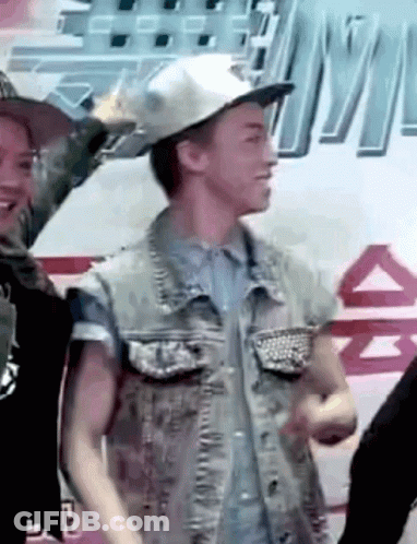 A Man Wearing A Hat And A Denim Vest Is Standing In Front Of A Sign That Says ' A ' On It GIF
