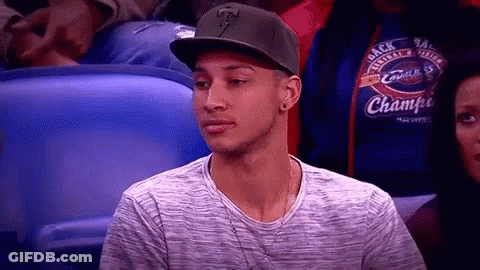 A Man Wearing A Hat And A Gray Shirt Is Sitting In A Stadium Watching A Game . GIF
