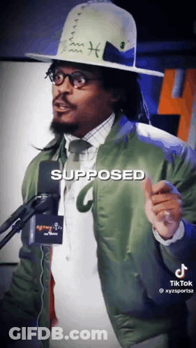 A Man Wearing A Hat And A Green Jacket Is Standing In Front Of A Microphone With The Word Supposed Written On It GIF
