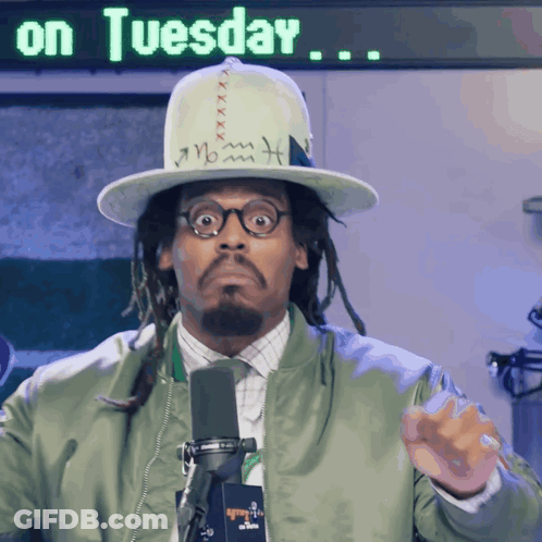 A Man Wearing A Hat And A Green Jacket Stands In Front Of A Sign That Says " On Tuesday " GIF