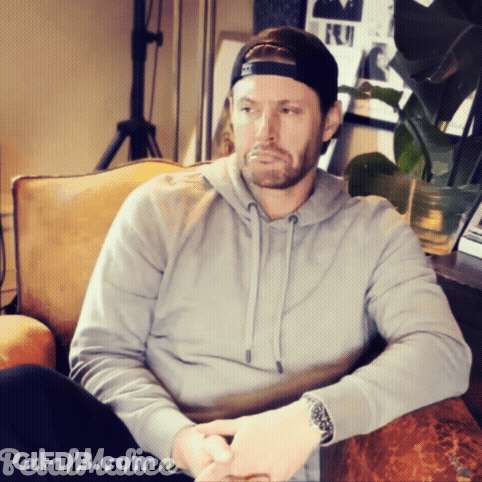 A Man Wearing A Hat And A Grey Hoodie Is Sitting On A Couch With Petal Malice Written On The Bottom GIF