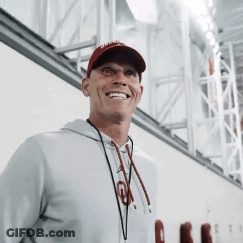 A Man Wearing A Hat And A Hoodie Is Smiling While Standing In A Gym . GIF