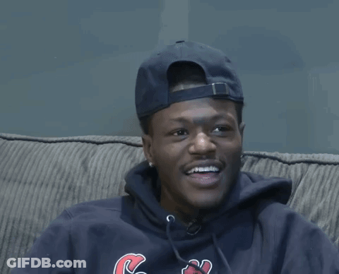 A Man Wearing A Hat And A Hoodie With The Letter C On It Sits On A Couch GIF