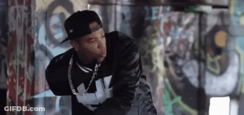 A Man Wearing A Hat And A Leather Jacket Is Standing In Front Of A Graffiti Wall . GIF