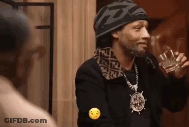 A Man Wearing A Hat And A Necklace Is Holding A Glass Of Alcohol . GIF