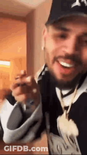A Man Wearing A Hat And A Necklace Is Holding A Pair Of Shoes . GIF