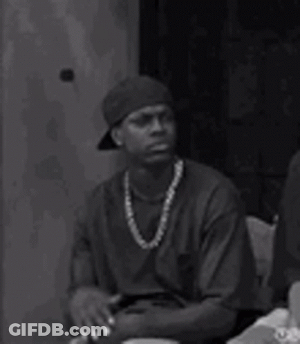 A Man Wearing A Hat And A Necklace Is Sitting At A Table With His Hands Folded . GIF