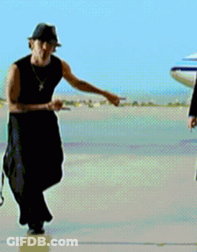 A Man Wearing A Hat And A Necklace Is Standing In Front Of An Airplane GIF