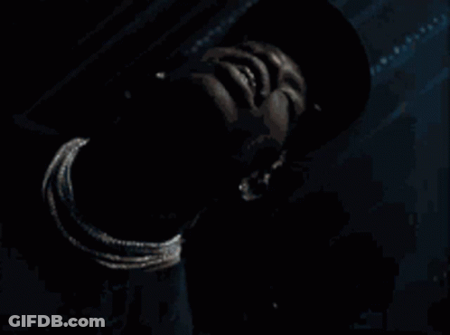A Man Wearing A Hat And A Necklace Looks Down At Something GIF