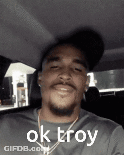A Man Wearing A Hat And A Necklace Says Ok Troy On His Face GIF