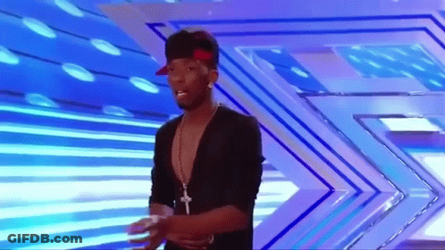 A Man Wearing A Hat And A Necklace Stands On A Stage GIF
