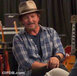 A Man Wearing A Hat And A Plaid Shirt Is Sitting In Front Of Guitars GIF