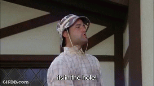 A Man Wearing A Hat And A Plaid Shirt Is Standing In Front Of A Building And Saying `` Its In The Hole '' . GIF