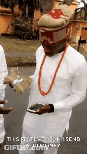 A Man Wearing A Hat And A Red Necklace Is Holding A Cell Phone And A Bag Of Money . GIF