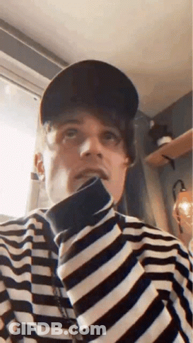 A Man Wearing A Hat And A Striped Shirt Is Looking Up At The Camera . GIF