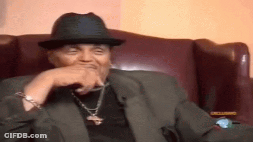 A Man Wearing A Hat And A Suit Is Sitting In A Chair With His Hand On His Chin . GIF