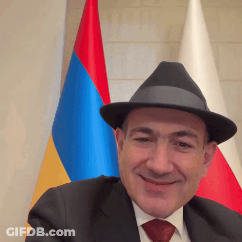 A Man Wearing A Hat And A Suit Smiles In Front Of A Flag GIF