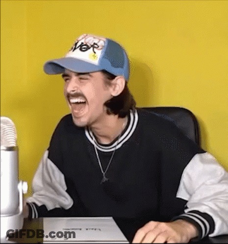A Man Wearing A Hat And A Sweater Is Sitting At A Desk Laughing . GIF