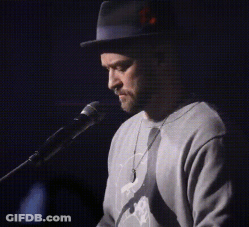 A Man Wearing A Hat And A Sweater Singing Into A Microphone GIF