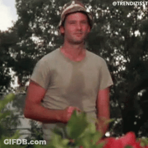 A Man Wearing A Hat And A T-Shirt Is Standing Next To A Plant . GIF