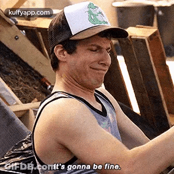 A Man Wearing A Hat And A Tank Top Is Sitting In Front Of A Pile Of Wooden Crates . GIF