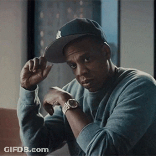 A Man Wearing A Hat And A Watch Is Sitting At A Table With His Hand On His Hat . GIF