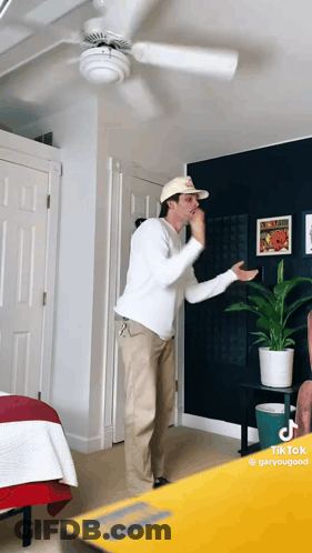 A Man Wearing A Hat And A White Shirt Is Standing In A Room With A Ceiling Fan GIF